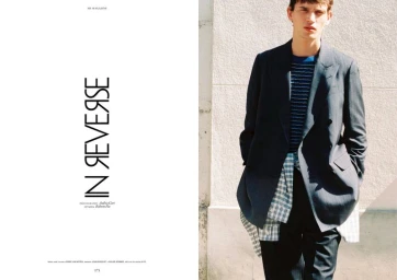 Magazine editorial spread titled In Reverse showing a male model in a dark blazer and layered plaid shirt against a sunlit wall.