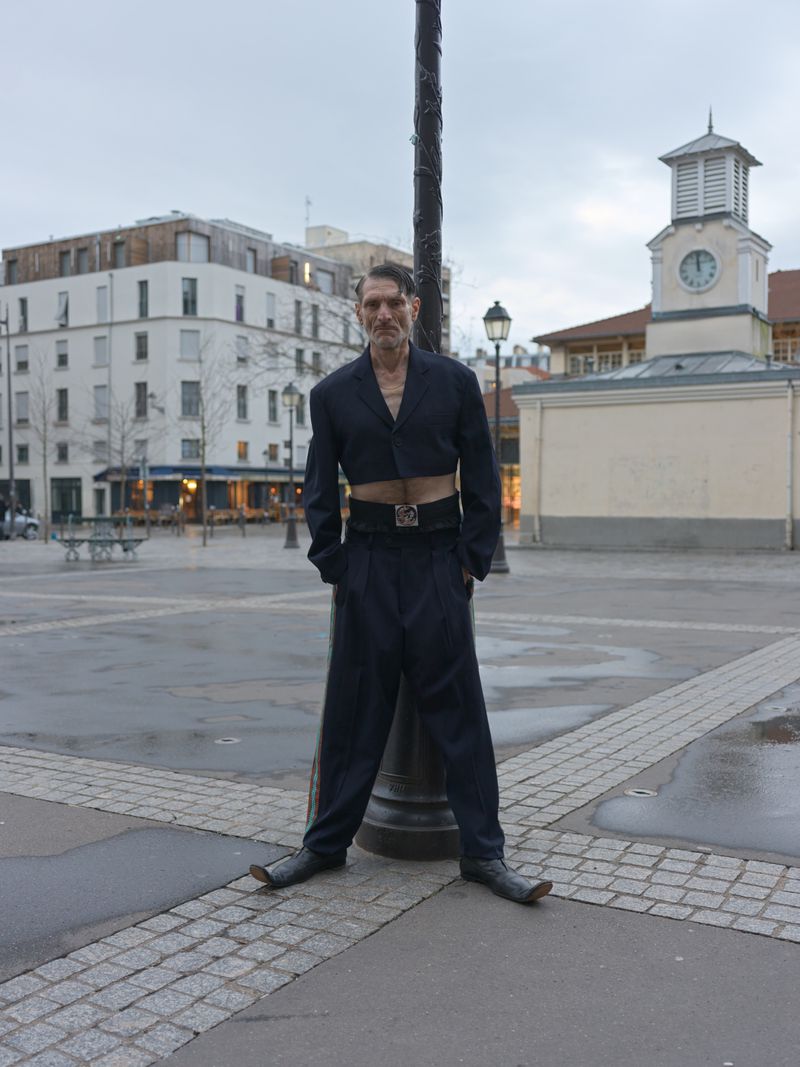 Adult male in cropped suit jacket and wide-legged trousers poses outdoors in front of a square with buildings and a clock tower.
