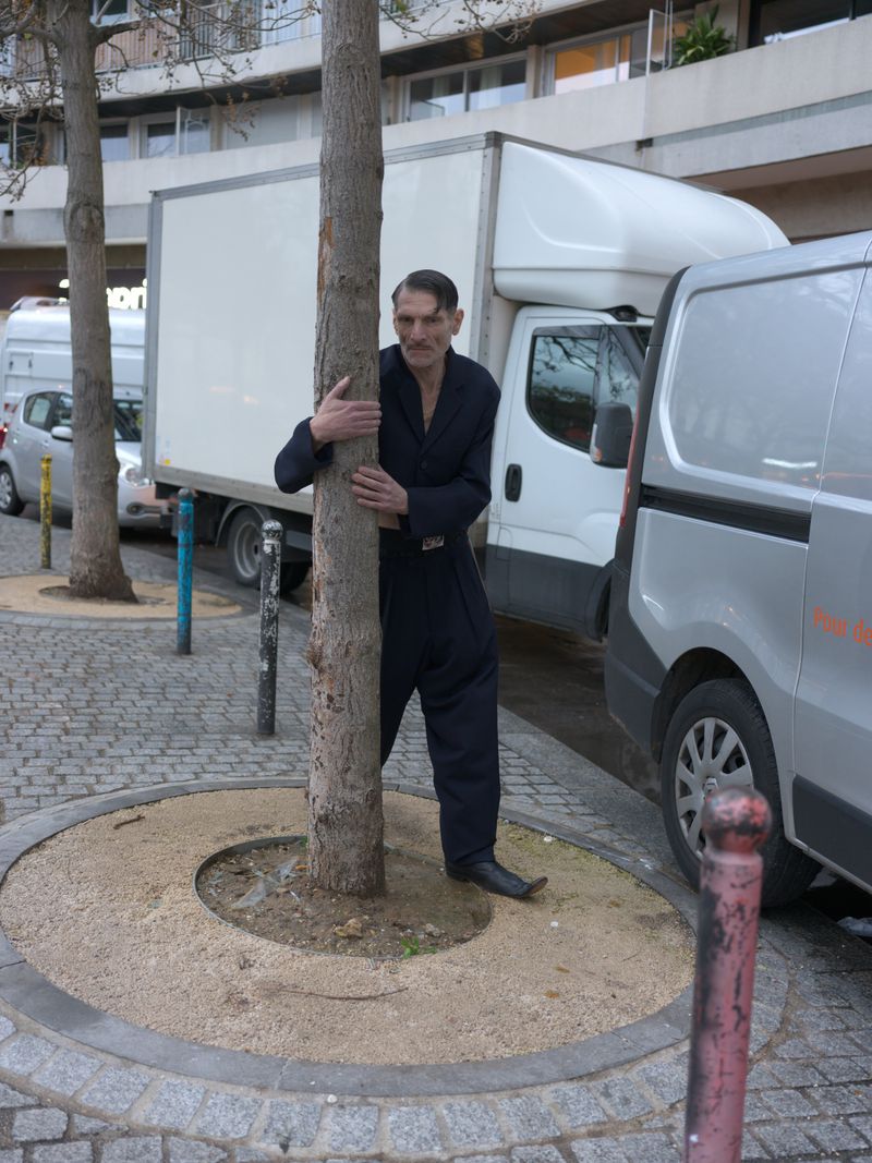 A man in a dark jumpsuit hugs a tree on a city street, with delivery trucks parked nearby in the background, on a cloudy day.