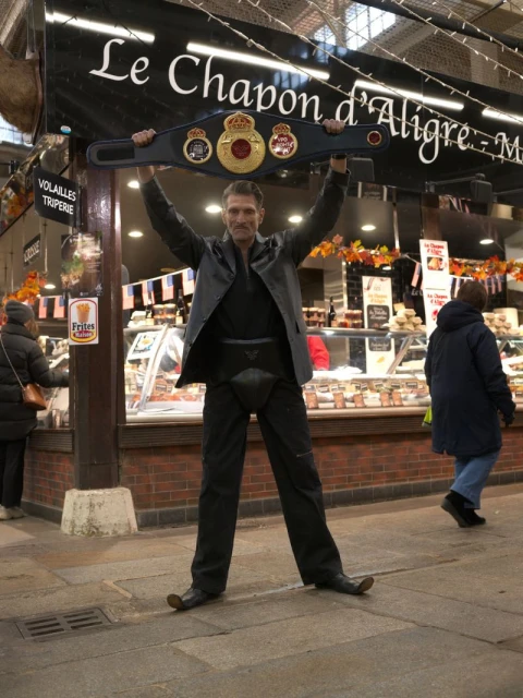 Man holds a boxing champion belt above his head, standing in front of Le Chapon d'Aligre store, wearing a black leather jacket.