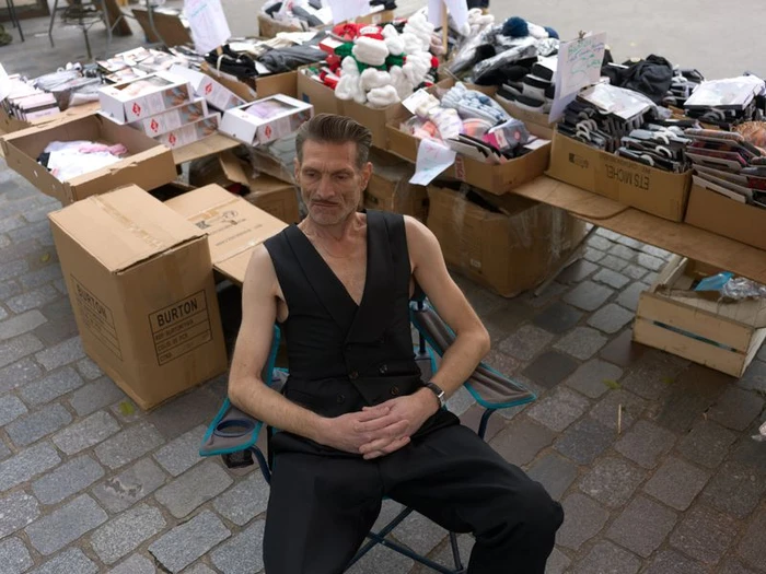 Man wearing a black vest and trousers sitting in a folding chair outdoors surrounded by market boxes
