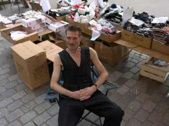 Man wearing a black vest and trousers sitting in a folding chair outdoors surrounded by market boxes