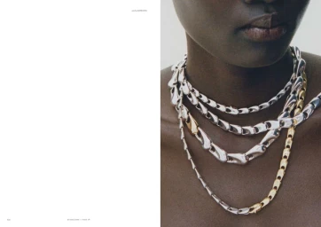 Close-up portrait of a Black woman wearing layered sculptural silver and gold chain necklaces on an editorial magazine spread.