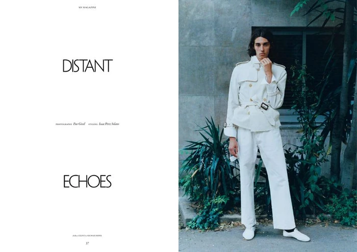 Male model in an all-white belted jacket, trousers, and white shoes posing outdoors next to green foliage for a magazine spread.