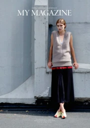 Model Anna Robinson posing against a grey concrete wall in a beige sleeveless knit sweater, red belt, and black skirt.