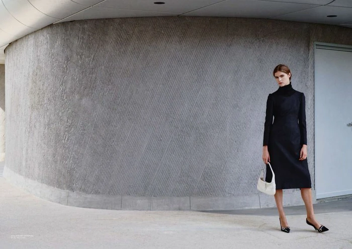 Model Anna Robinson wearing a long-sleeve black midi dress and holding a white handbag against a curved textured concrete wall.