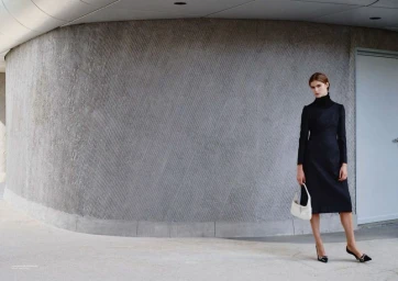 Model Anna Robinson wearing a long-sleeve black midi dress and holding a white handbag against a curved textured concrete wall.
