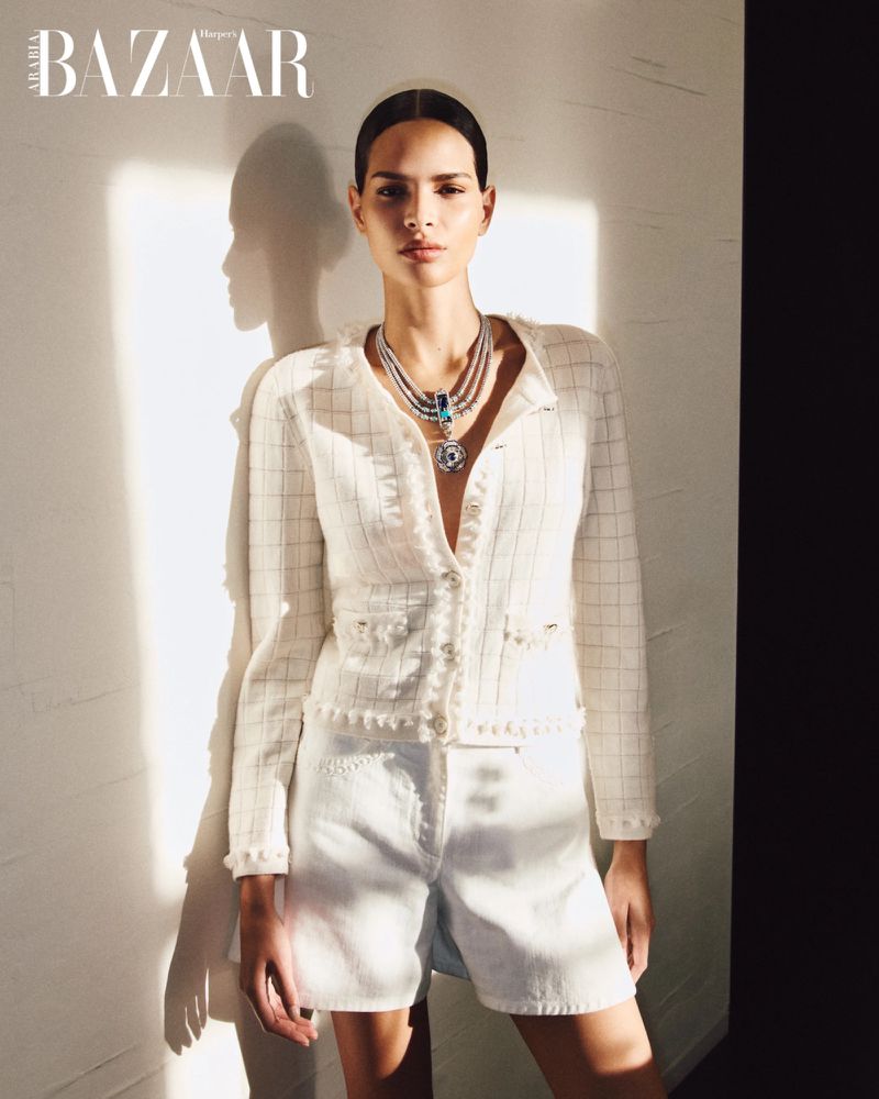 Model wearing a white tweed jacket, matching shorts, and a layered blue statement necklace against a sunlit wall.