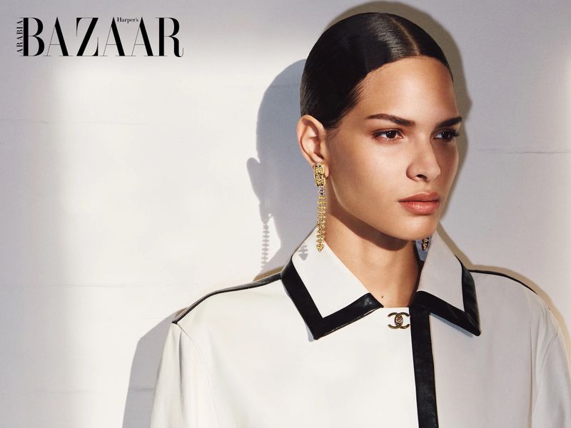 Model with slicked-back hair wearing dangling gold earrings and a white jacket with black trim against a white background.