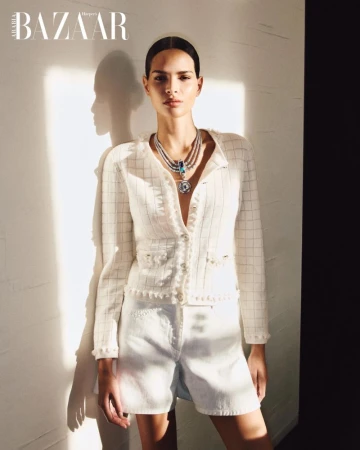 Model wearing a white tweed jacket, matching shorts, and a layered blue statement necklace against a sunlit wall.