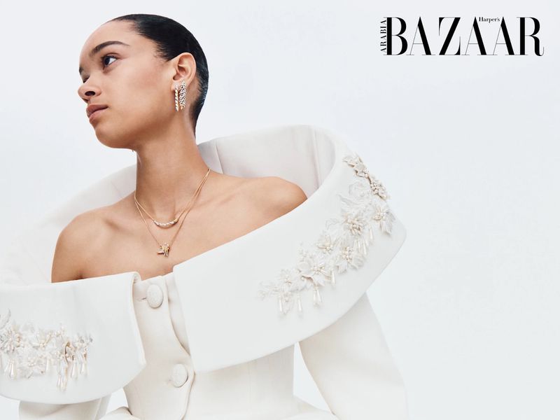 Model wearing an architectural white off-shoulder coat with floral beadwork and gold jewelry against a white studio background.
