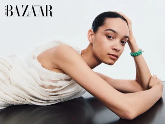 Woman in a textured off-white layered dress leaning on a dark reflective surface wearing a green bracelet against a white background.