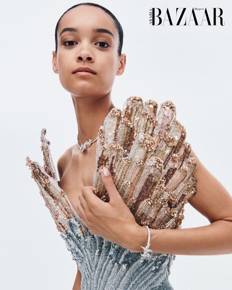 Close-up portrait of a woman in an ornate beaded sculptural gown wearing diamond jewelry against a light studio backdrop.