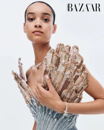 Close-up portrait of a woman in an ornate beaded sculptural gown wearing diamond jewelry against a light studio backdrop.