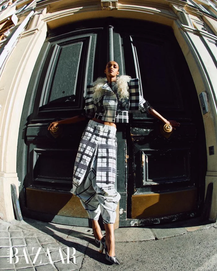 Model Muna Mahamed in a black and white plaid suit posing against large dark doors for Harper's Bazaar Arabia.