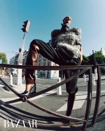 Model Muna Mahamed posing behind a metal street guardrail wearing a fuzzy high-neck top and leather boots in an urban street.