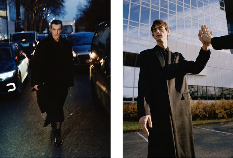 Two adult men in dark overcoats are captured in an outdoor editorial fashion context, with urban and architectural backgrounds. 
