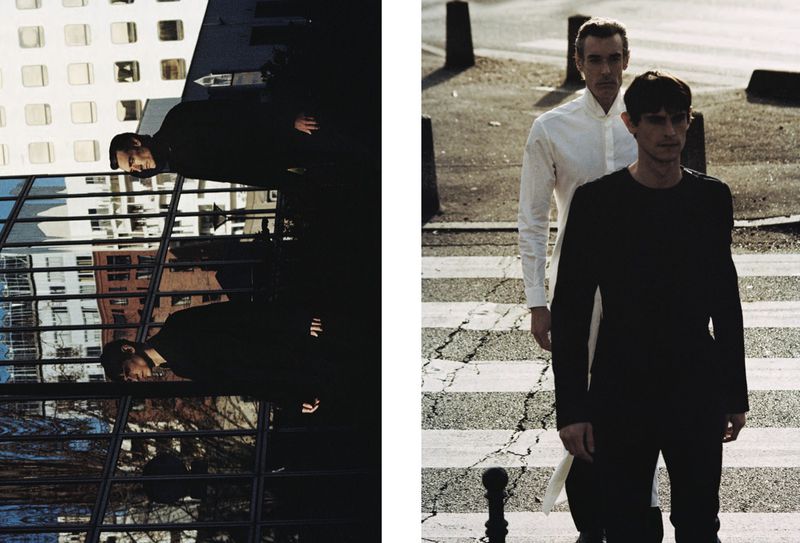 Two adult men stand on a crosswalk in an urban environment; on the left, an office building reflects a man in a suit.