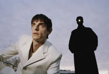 Man wearing a white jacket looking upward with a silhouetted figure standing in the background under a pale sky