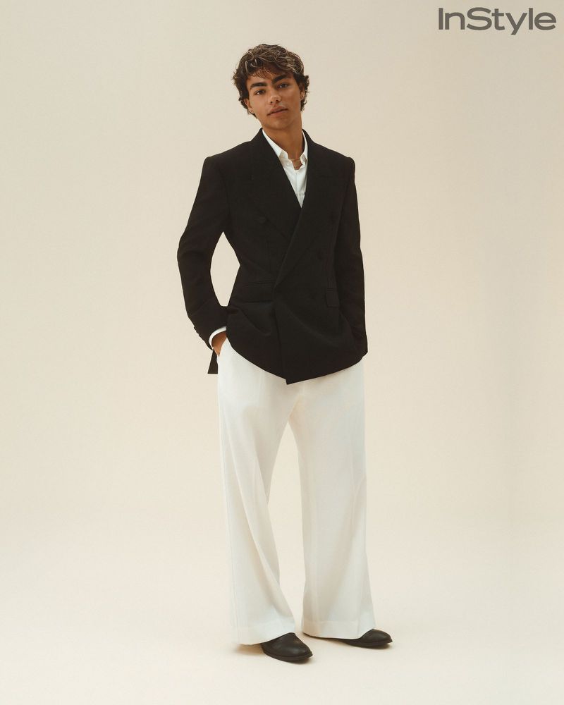 A young man wearing a black double-breasted blazer and wide-leg white trousers poses against an off-white seamless studio backdrop.