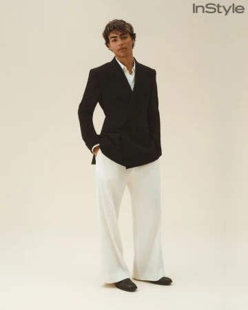 A young man wearing a black double-breasted blazer and wide-leg white trousers poses against an off-white seamless studio backdrop.