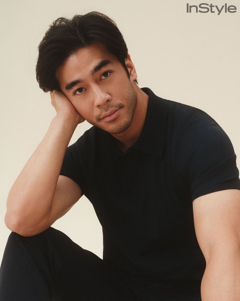 Portrait of actor Alex Landi wearing a black short-sleeved polo shirt resting his head on his hand against a neutral studio backdrop.
