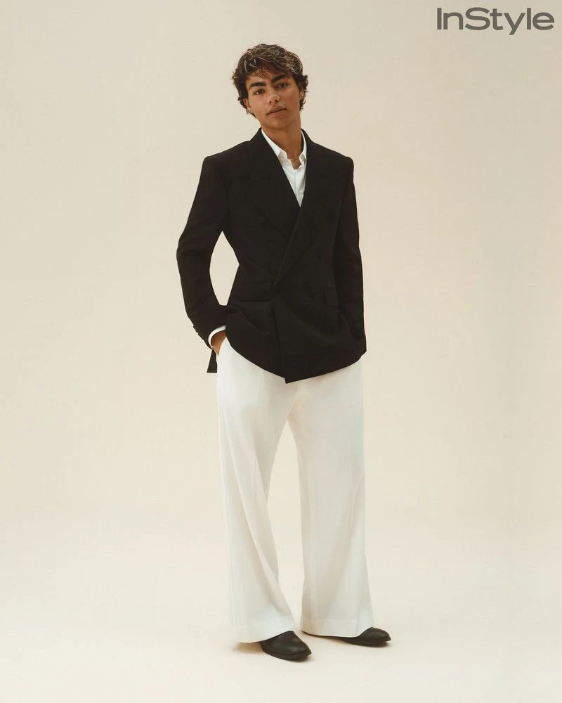 A young man wearing a black double-breasted blazer and wide-leg white trousers poses against an off-white seamless studio backdrop.
