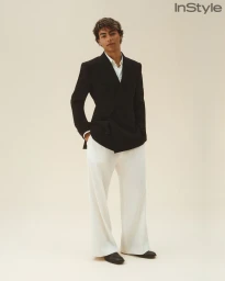 A young man wearing a black double-breasted blazer and wide-leg white trousers poses against an off-white seamless studio backdrop.