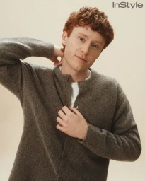 Young man with curly red hair wearing a grey knit cardigan button-down over a white shirt against a neutral cream studio backdrop.