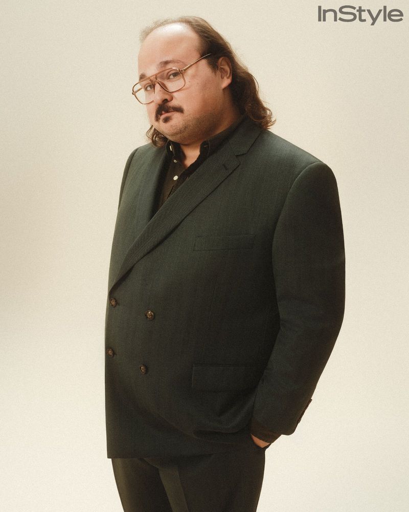 Stavros Halkias in a dark double-breasted blazer and wire-rim glasses standing against a warm studio backdrop.