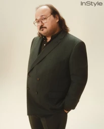 Stavros Halkias in a dark double-breasted blazer and wire-rim glasses standing against a warm studio backdrop.