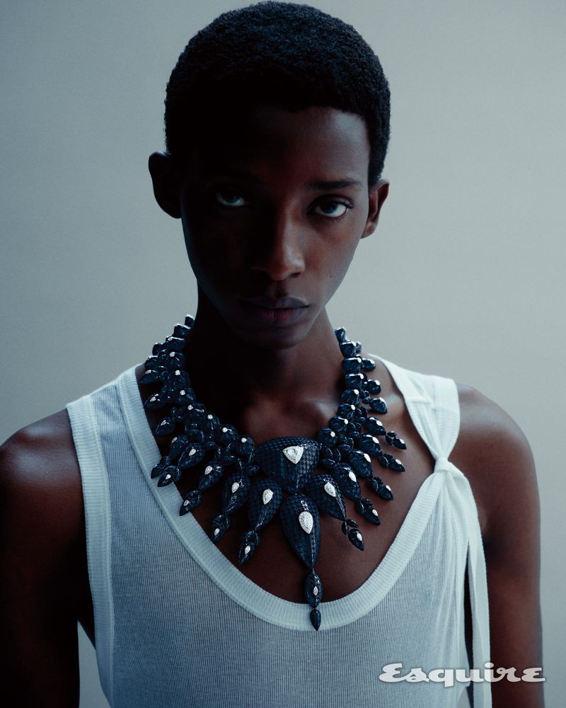 Gideon Adeniyi wearing an ornate black statement necklace and a white ribbed tank top against a pale grey studio background.