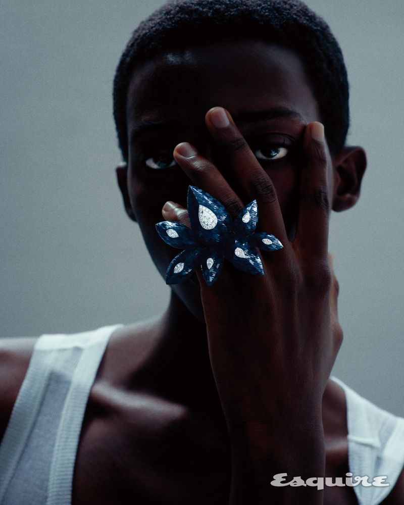 Close-up of a Black man wearing a white ribbed tank top holding an ornate blue gemstone ring in front of his face against a grey backdrop.