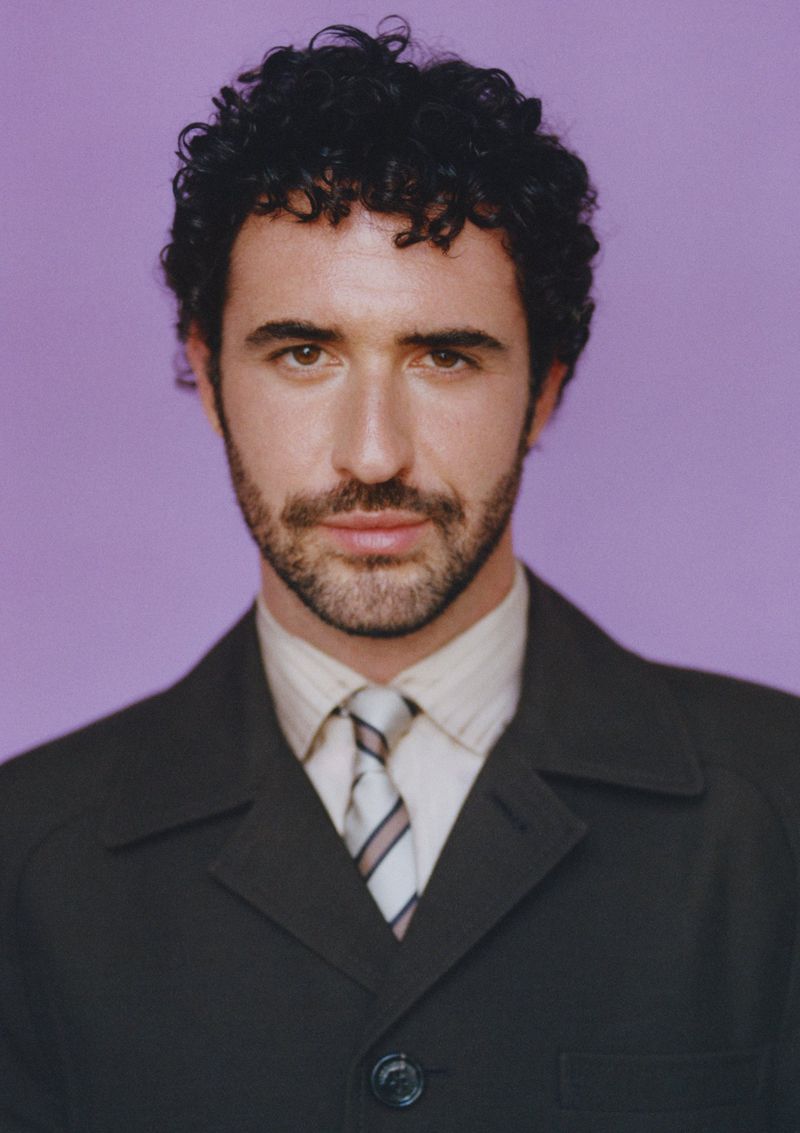 Jordan Firstman wearing a dark suit jacket, collared shirt, and striped tie against a solid light purple studio backdrop.