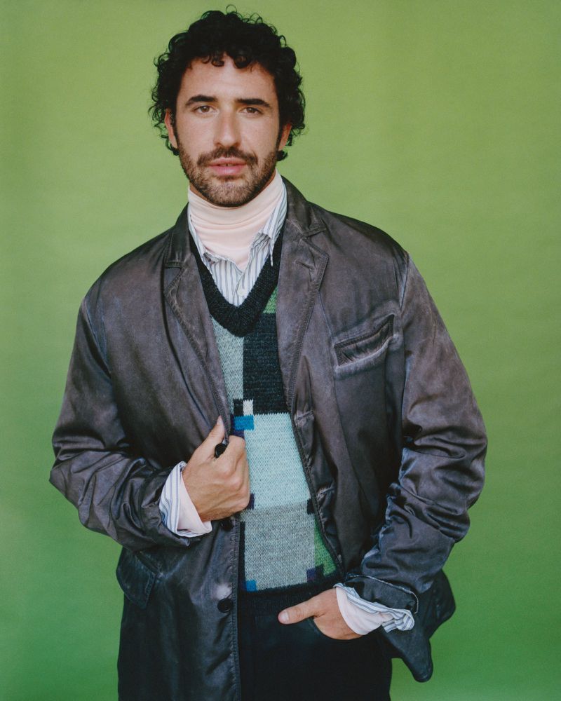 Jordan Firstman wearing a dark layered jacket and patterned knit vest against a solid green background.