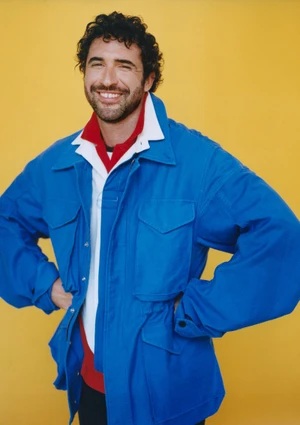 Jordan Firstman smiling in an oversized blue utility jacket over layered collared shirts against a solid yellow backdrop.