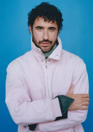 Jordan Firstman posing with arms crossed in a pale pink high-collar jacket against a solid blue background.