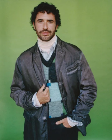Jordan Firstman wearing a dark layered jacket and patterned knit vest against a solid green background.
