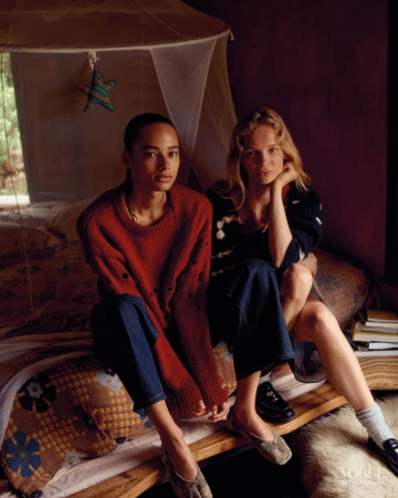 Two models sitting side by side on a rustic bed wearing knit sweaters and dark denim in a moody bedroom setting.