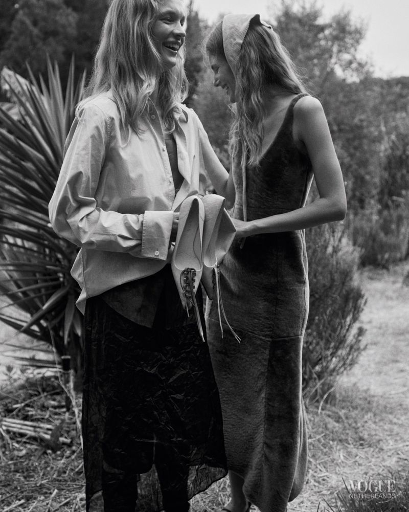 Two women laughing outdoors in black and white, one wearing an oversized shirt and dark skirt holding shoes, the other in a slip dress.