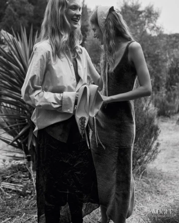 Two women laughing outdoors in black and white, one wearing an oversized shirt and dark skirt holding shoes, the other in a slip dress.