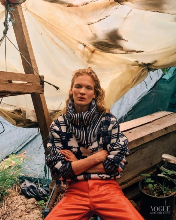Model with crossed arms wearing a patterned turtleneck knit sweater and bright red-orange trousers inside a makeshift tarp shelter.