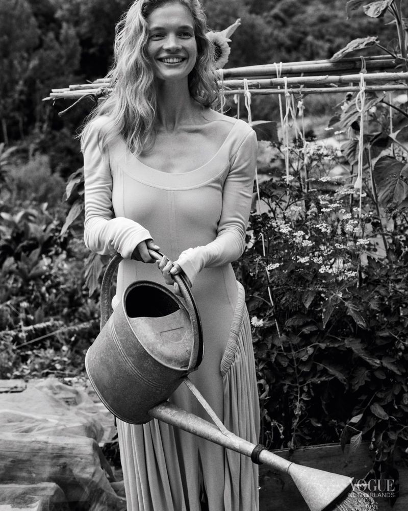 Smiling woman wearing a long-sleeve scoop neck dress holding a large metal watering can in an outdoor garden.