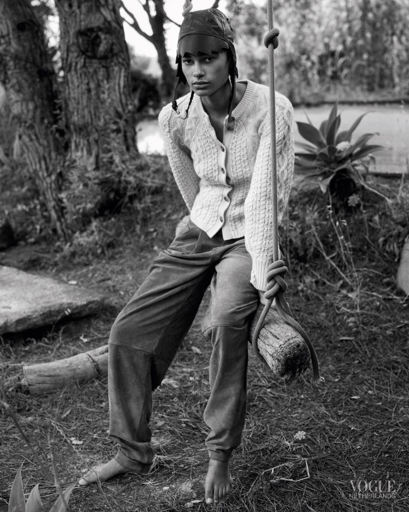 Model wearing a cable-knit cardigan, loose trousers, and leather cap sitting barefoot on a wooden rope swing outdoors.