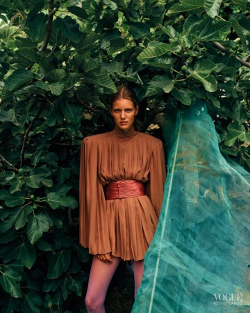 Model wearing a pleated terracotta mini dress with wide sash and pink tights posed before lush green fig leaves and draped teal netting.