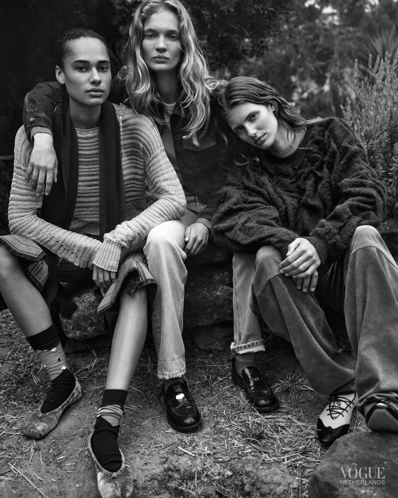 Three female models seated closely together on a rocky ground outdoors wearing textured knitwear for Vogue Netherlands.