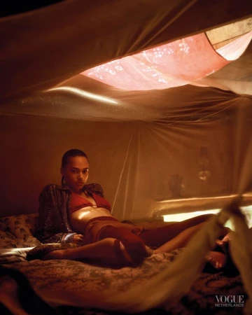 Model reclining inside a sheer draped tent wearing a red bikini top and patterned jacket in warm atmospheric light.
