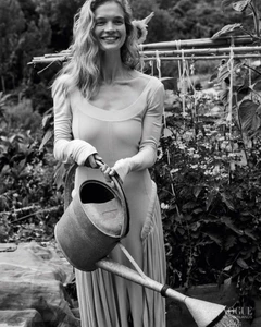 Smiling woman in a long-sleeve dress holding a vintage metal watering can in an outdoor vegetable garden in black and white.