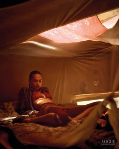 Model reclining in a red bikini top and patterned shirt inside a warm, backlit fabric canopy.