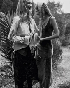 Two women laughing outdoors in black and white, one wearing an oversized shirt and dark skirt holding shoes, the other in a slip dress.